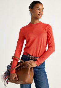 Rød langærmet sweater med ribbet halsudskæring og knapdetaljer, parret med en brun lædertaske, hvor en mønstret tørklæde er bundet til håndtaget.