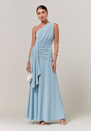 Frau trägt ein hellblaues One-Shoulder-Kleid mit Drapierung, hält eine weiße Clutch in Muschelform und ist mit goldenen Schmuckstücken und Sandalen ausgestattet.