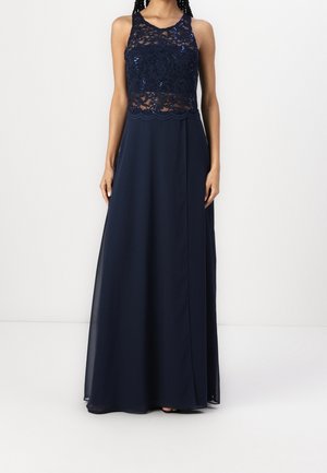 Femme portant une robe de soirée bleu marine sans manches avec un corsage en dentelle et sequins et une longue jupe fluide.