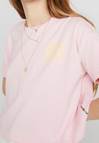Žena v růžovém tričku s žlutým logem "Home Boy" na hrudi a malou duhovou nášivkou na postranním švu, doplněná o vrstvené zlaté náhrdelníky.