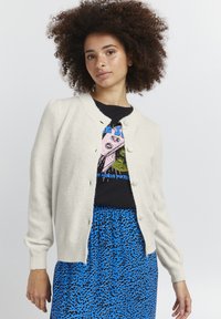 Cárdigan crema con cuello redondo y cinco botones. Textura suave, mangas largas, usado sobre una camiseta gráfica negra y una falda de estampado de leopardo azul.