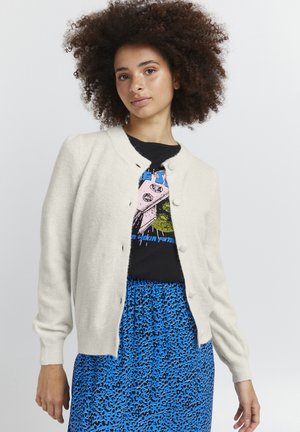 Cremefarbener Cardigan mit Rundhalsausschnitt und fünf Knopfverschlüssen. Weicher Stoff, lange Ärmel, über einem schwarzen Grafik-T-Shirt und einem blauen Leopardenrucksack getragen.