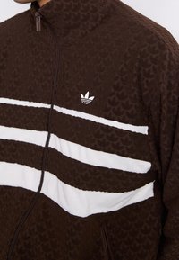 Ruskea vetoketjullinen takki, jossa on tekstuuri, kolme valkoista vaakasuoraa raitaa ja pieni valkoinen logo rinnassa.