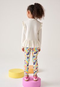 Witte top met lange mouwen en ruches aan de mouwen, peplumzoom; meerkleurige leggings met regenboog- en sterrenpatroon; roze clogs; staand op kleurrijke podia.