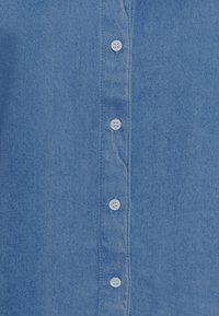 Světle modrá denimová košile s knoflíkovým zapínáním; má bílé knoflíky a hladkou texturu, která zvýrazňuje detaily švů.