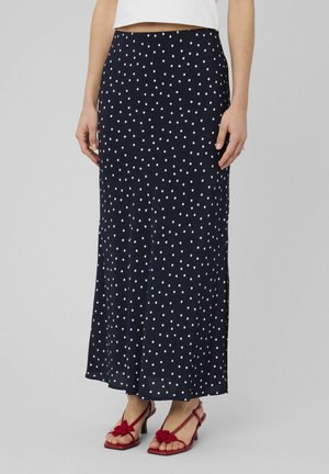 Femme portant une jupe longue bleu marine à pois blancs, associée à des sandales rouges à talons ouvertes et un top blanc court.