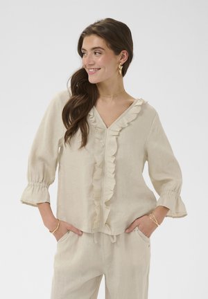 Femeie cu păr lung și castaniu, purtând o bluză bej cu volane și pantaloni asortați, zâmbind și privind în lateral, cu mâinile în buzunare.