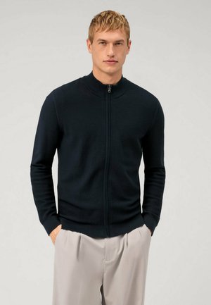 Navy ribbed knit jacket with a stand collar and a front zipper, featuring long sleeves and a fitted silhouette, paired with light trousers.