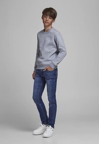 Ragazzo adolescente in piedi con una mano in tasca, indossa un maglione grigio chiaro, jeans blu e scarpe da ginnastica bianche su uno sfondo grigio uniforme.