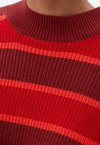 Strickpullover mit breiten horizontalen Streifen in Dunkelrot, Hellrot und Orange. Der Kragen ist gerippt und hochgeschlossen.