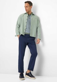 Lichtgroene linnen shirt over een blauw patroon polo, gecombineerd met marineblauwe chino's en donkere sneakers. Het shirt heeft een borstzak en knoopsluiting.