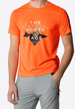 T-shirt orange vif à manches courtes avec le logo "The North Face" et un graphique abstrait de montagne, porté avec un pantalon gris clair.