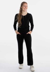 Zwarte lange mouwen top gecombineerd met zwarte joggingsbroek met elastische tailleband en twee voorkeurszakken. Model draagt witte sneakers.