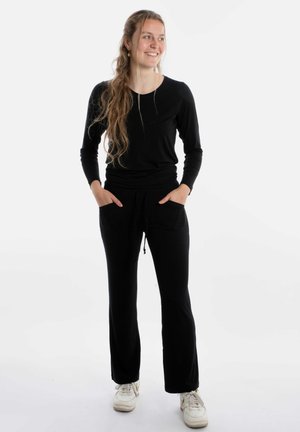 Zwarte lange mouwen top gecombineerd met zwarte joggingsbroek met elastische tailleband en twee voorkeurszakken. Model draagt witte sneakers.