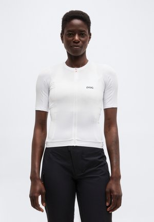 Άτομο που φοράει στενή λευκή κοντομάνικη μπλούζα με φερμουάρ και λογότυπο "poc" και μαύρο παντελόνι, στέκεται μπροστά σε απλό φωτεινό φόντο.