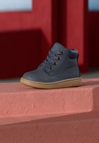 Bota de cuero gris hasta el tobillo con punta redonda, suela de goma contrastante, ojales dorados y cordones grises. Acabado suave con un cuello acolchado.
