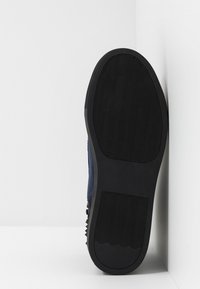 Czarny gumowy bieżnik z teksturowaną, falistą powierzchnią. Profil boczny ukazuje ciemny materiał sięgający górnej części buta. Czysty design, minimalne detale.