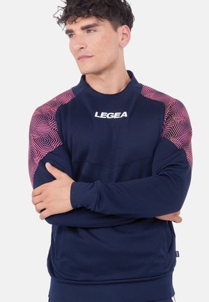 Uomo con capelli ricci e scuri che indossa una maglia sportiva a maniche lunghe blu navy con motivi geometrici rosa sulle spalle e logo "LEGEA" sul petto.