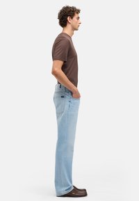 Jasnoniebieskie dżinsy o kroju prostych nogawek, zestawione z brązowym t-shirtem z krótkim rękawem. Model stoi bokiem, ma na sobie brązowe buty.