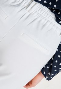 Witte broek gemaakt van zachte stof, met een elastische tailleband en een flatte achterzak. Gecombineerd met een marineblauw shirt met witte hartjespatronen.