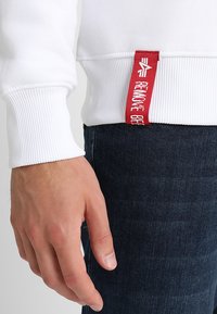 Hvid sweatshirt med ribbede manchetter; har et rødt stofmærke med hvid tekst. Model iført mørke jeans; nærbillede af hånd og ærme.