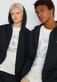 Des sweat-shirts à capuche zippés noirs en tissu doux, portés sur des t-shirts clairs avec des textes graphiques. Ils présentent une coupe décontractée et des poches frontales.