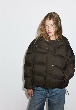 Junge Frau mit langen Haaren, die eine dunkle Steppjacke und blaue Jeans trägt, vor einem schlichten weißen Hintergrund stehend.
