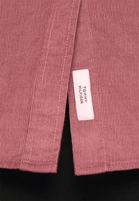 Roze corduroystof met een geribbeld textuur, voorzien van een klein wit label met de tekst "TOMMY HILFIGER" op de naad.