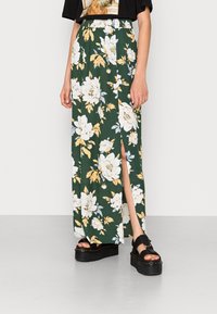 Jupe maxi florale en tissu vert avec des fleurs blanches et jaunes, dotée d'une fente latérale. Assortie à des sandales à plateforme noires.