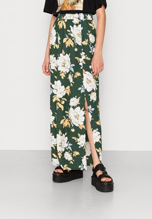 Jupe maxi florale en tissu vert avec des fleurs blanches et jaunes, dotée d'une fente latérale. Assortie à des sandales à plateforme noires.