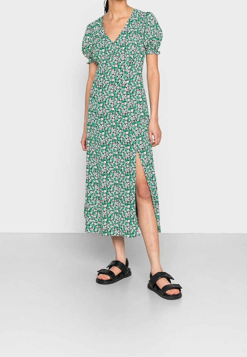 Zelené květinové šaty s krátkými balónovými rukávy, výstřihem do V a rozparkem až ke stehnům. Hladká tkanina s přiléhavým korzetem a volnou sukní. Černé sandály.
