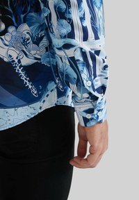 Chemise à manches courtes avec un motif floral abstrait bleu. Fabriquée en tissu léger. Comprend un poignet à boutons et un ourlet incurvé.