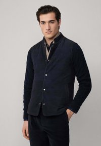 Mornaricki preklopni prsluk s visokim ovratnikom, srebrnim kopčama na dugmad i teksturiranim pletenim rukavima. Uparen s tamnom košuljom i hlačama.