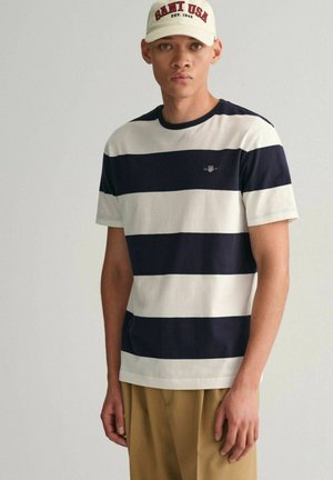 T-shirt à rayures bleu marine et blanches à manches courtes en coton, avec un col rond et un petit logo sur la poitrine. Associé à un pantalon beige.