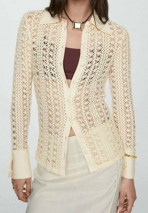Femme portant un chemisier en crochet crème boutonné sur un crop top marron et une jupe crème avec un collier pendentif carré et un bracelet en or.