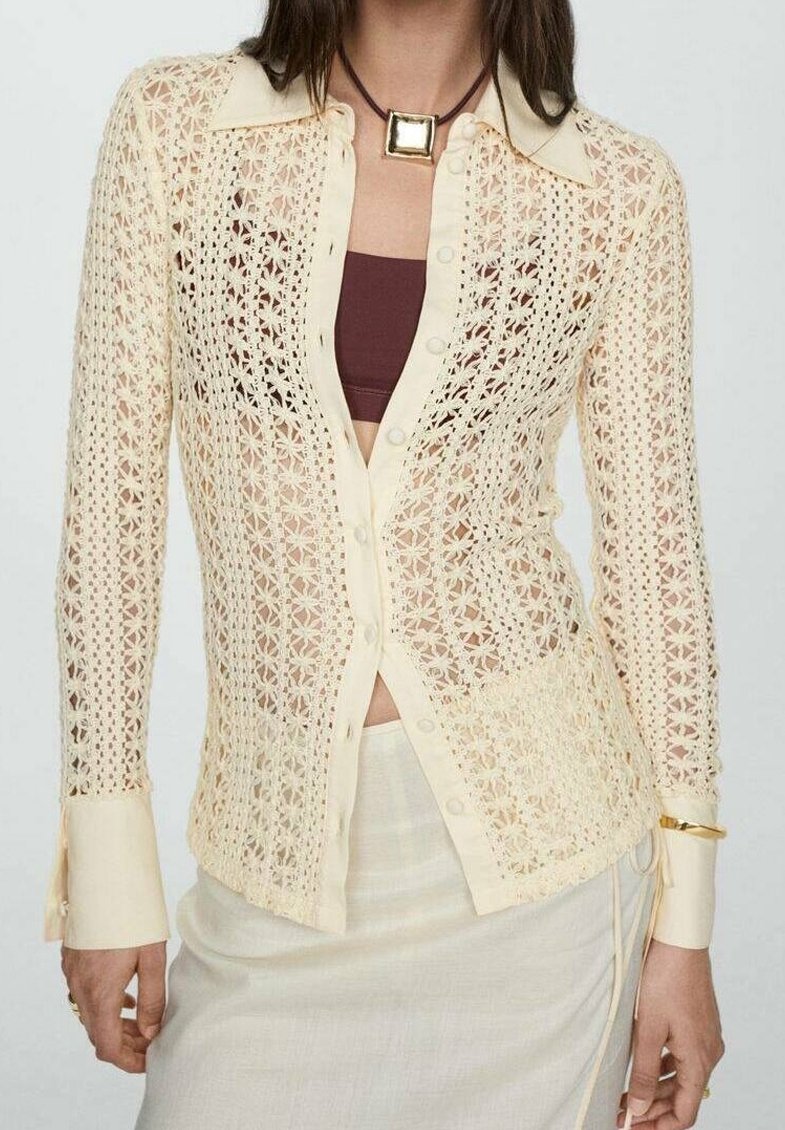 Femme portant un chemisier en crochet crème boutonné sur un crop top marron et une jupe crème avec un collier pendentif carré et un bracelet en or.