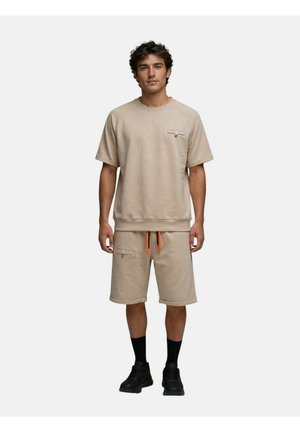 Jeune homme portant un sweat-shirt beige à manches courtes et un short assorti avec des cordons orange, des chaussettes noires et des baskets noires, debout sur fond blanc.