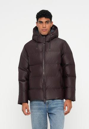 Junger Mann in einer dunkelbraunen gepolsterten Kapuzenjacke und hellblauen Jeans, der vor einem schlichten weißen Hintergrund steht.