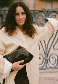 Camicia button-up a righe gialla, svasata, con polsini bianchi; tiene in mano una pochette nera in pelle texture su uno sfondo decorativo.