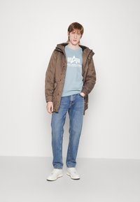 Giacca imbottita marrone con cappuccio sopra un maglione leggero blu chiaro con logo. Abbinata a jeans blu e sneakers bianche.