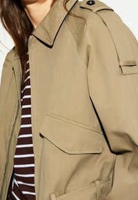 Khaki oversize bunda s velkými kapsami, ramenními epauletami a hladkým povrchem, doplněná hnědým a bílým pruhovaným tričkem.