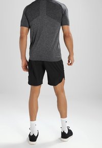 Pánský atletický outfit sestávající z šedého trička s krátkým rukávem a texturovanými rameny, doplněného černými šortkami. Oblečeno s bílými ponožkami a černými botami.