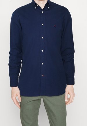 Homme portant une chemise bleu marine à manches longues boutonnée avec un petit logo et un pantalon vert olive, debout devant un fond uni.