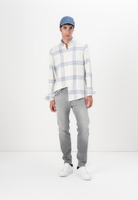 Rutete skjorte i hvit og lyseblå, grå jeans og hvite joggesko. Modellen har på seg en denimcaps og står foran en ensfarget bakgrunn.
