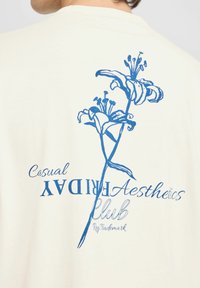 Cremefarvet t-shirt med et blåt blomstermotiv af liljer, med teksten "Casual Friday Aesthetics Club" i fede og kursiverede skrifttyper.