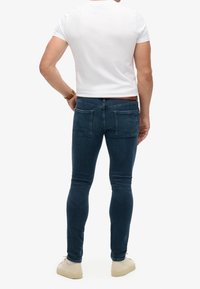 Jeans oscuros de hombre de corte slim con una textura suave y diseño de cinco bolsillos, combinados con una camiseta ajustada blanca y zapatillas beige.