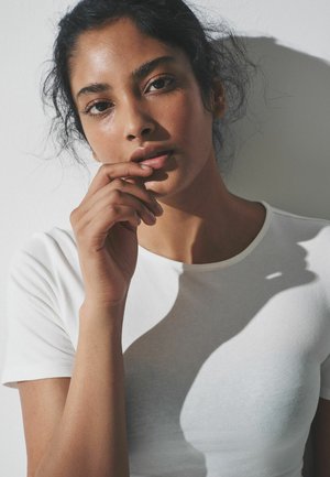 Valge lühikeste varrukatega t-särk, mida kannab inimene, millel on ümar kaelus ja sile kangas, naha peal tekitavad pehmed varjud kontraste.