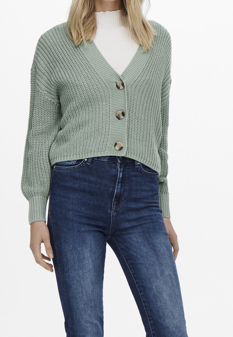 Femme portant un cardigan épais à grosse maille vert clair avec trois boutons, sur un haut blanc et un jean bleu foncé.