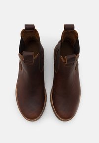 Καφέ δερμάτινα μποτάκια μέχρι τον αστράγαλο με ελαστικές πλαϊνές επιφάνειες και χερούλια. Διαθέτουν υφή και αντίθετη ανοιχτόχρωμη ραφή γύρω από τη σόλα.