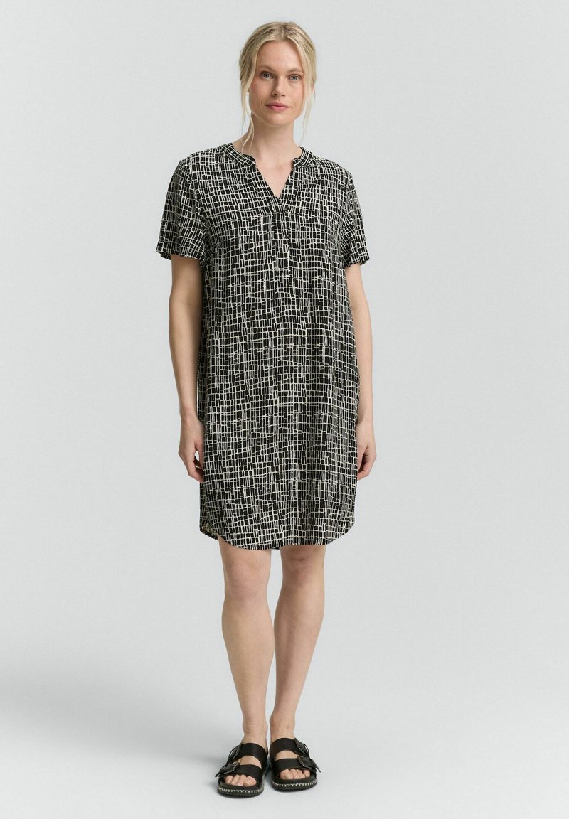 Robe noire et blanche à manches courtes avec un col en V, présentant un motif en carreaux, coupe décontractée, atteignant le milieu de la cuisse, associée à des sandales noires.